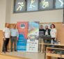 Group of five people standing on a stage next to banners for a charity event, indoors in a hall.